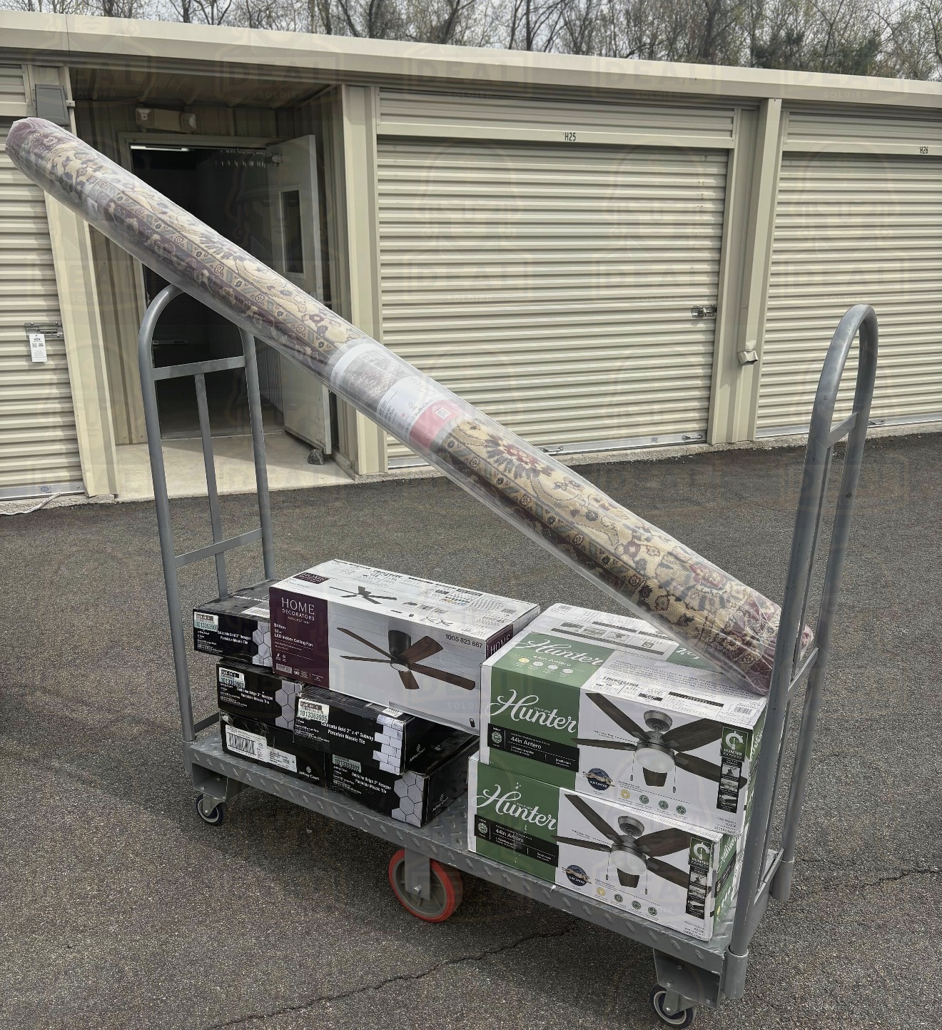 Home Depot orange flat cart parked outside a self-storage facility loaded with a rolled Marash Red 8 by 10 foot rug, three boxed Hunter and Home Decorators ceiling fans, and stacked tile boxes underneath.